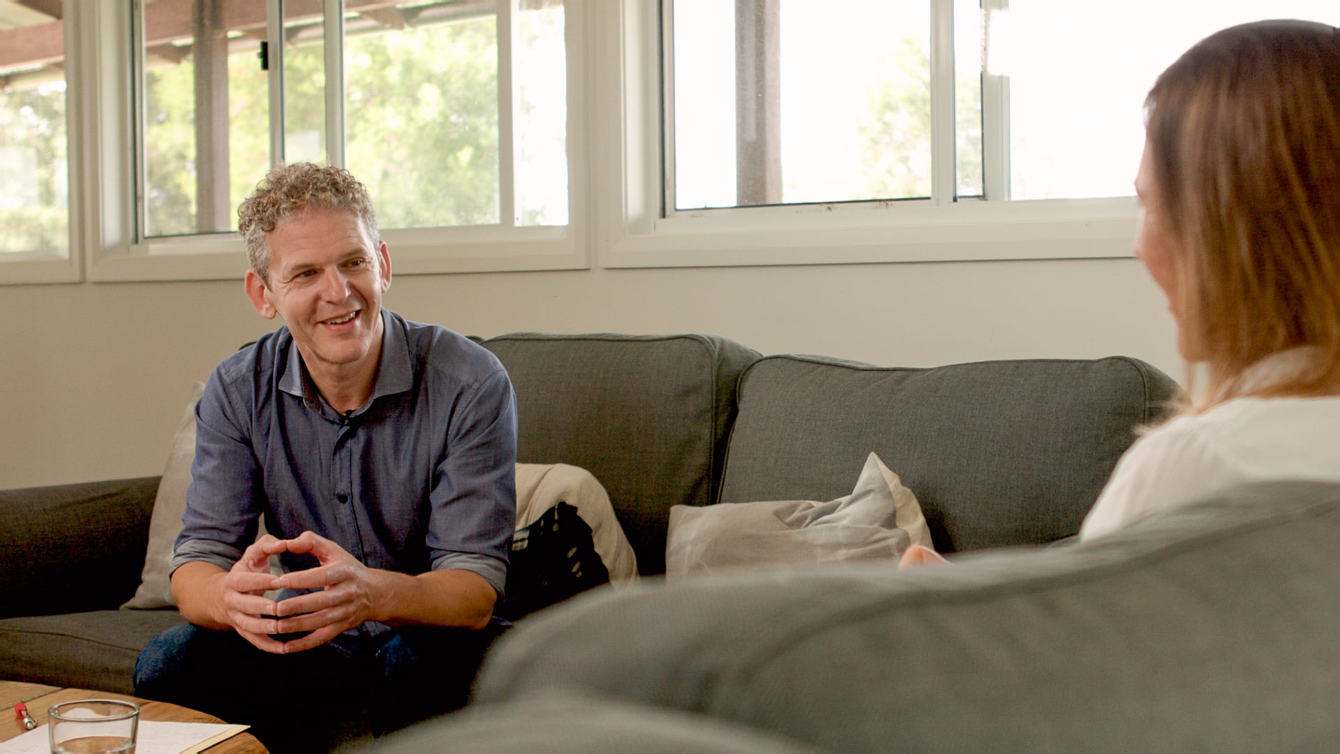 Russ sits on a couch, leaning forward with his hands clasped, speaking to a woman seated opposite him in a bright, comfortable room with large windows.