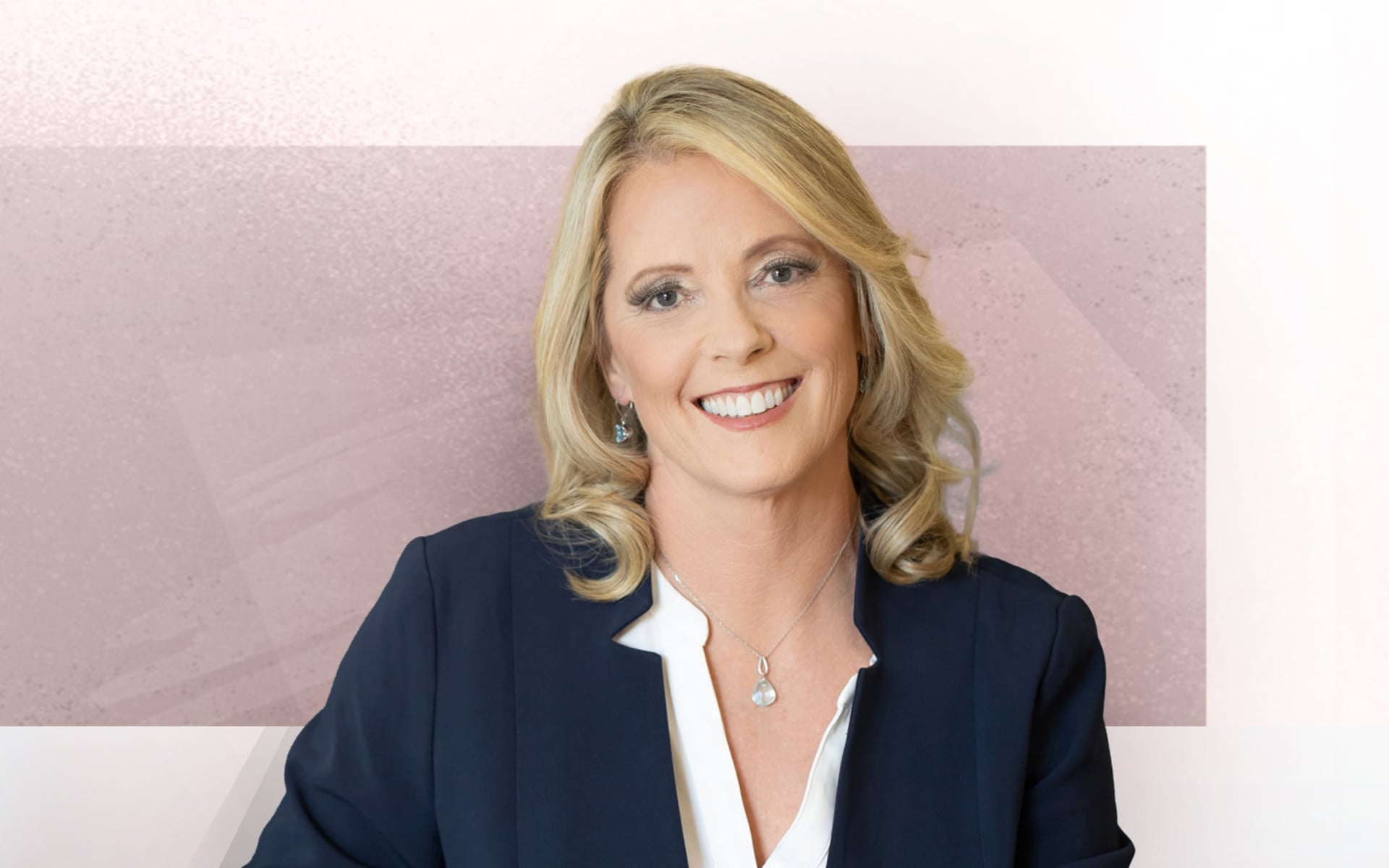 A woman(Dr Sara Gilman) with shoulder-length blonde hair, wearing a dark blazer and a white top, smiles at the camera. The background is a blend of muted pink and gray tones.
