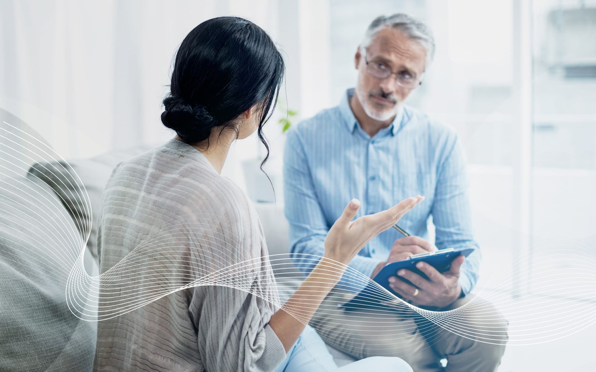 A woman with long dark hair is speaking, facing away from the camera, while a man with gray hair and glasses listens attentively and takes notes on a clipboard. The setting appears to be a bright indoor space, possibly a counseling or consultation environ