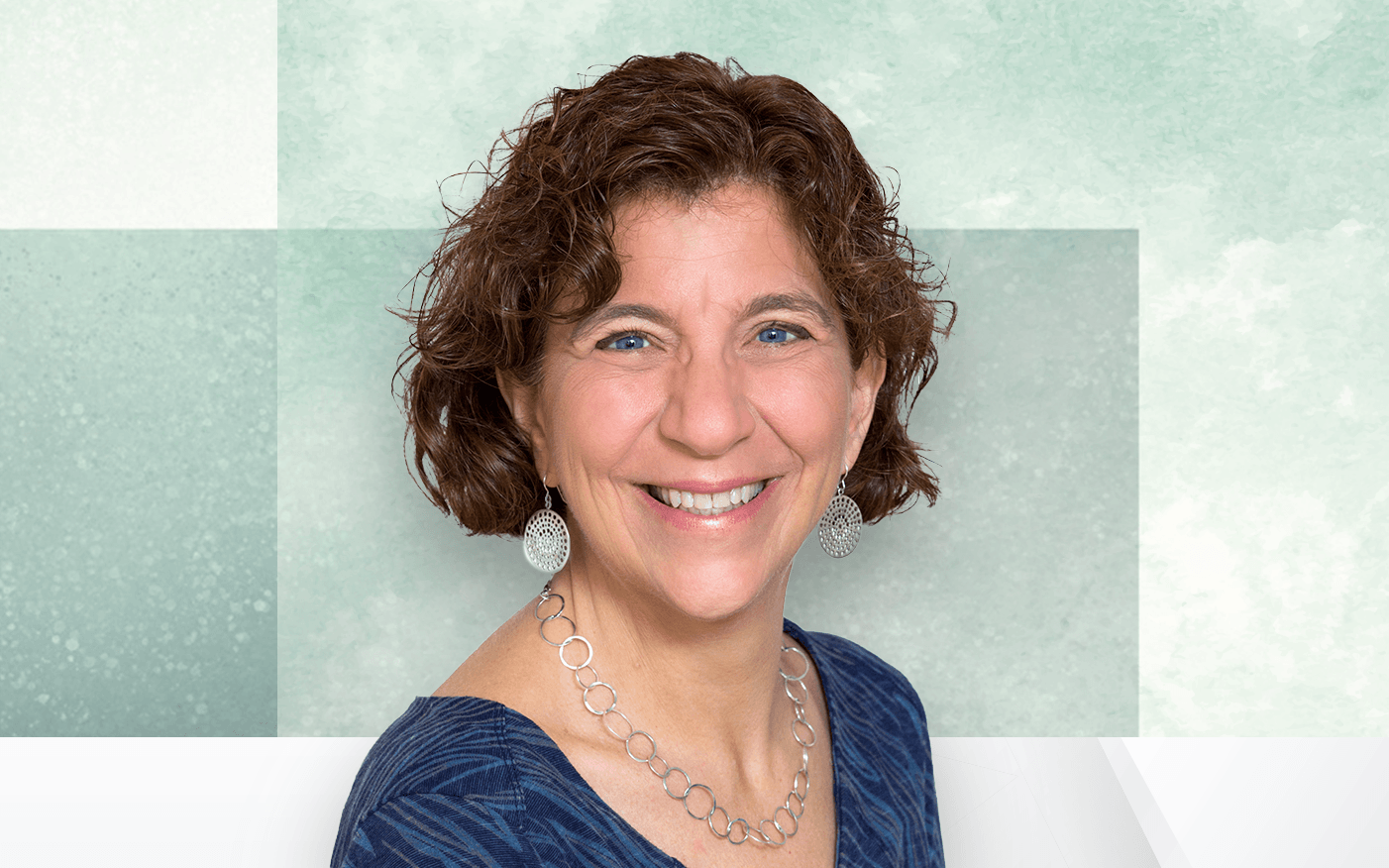 Deb Korn smiles at the camera, wearing silver earrings and a necklace. She is dressed in a blue patterned top. The background features soft shades of green and white geometric shapes.